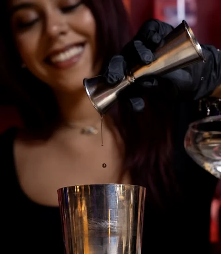 A bartender pouring from a jigger into a mixing tin under red light
