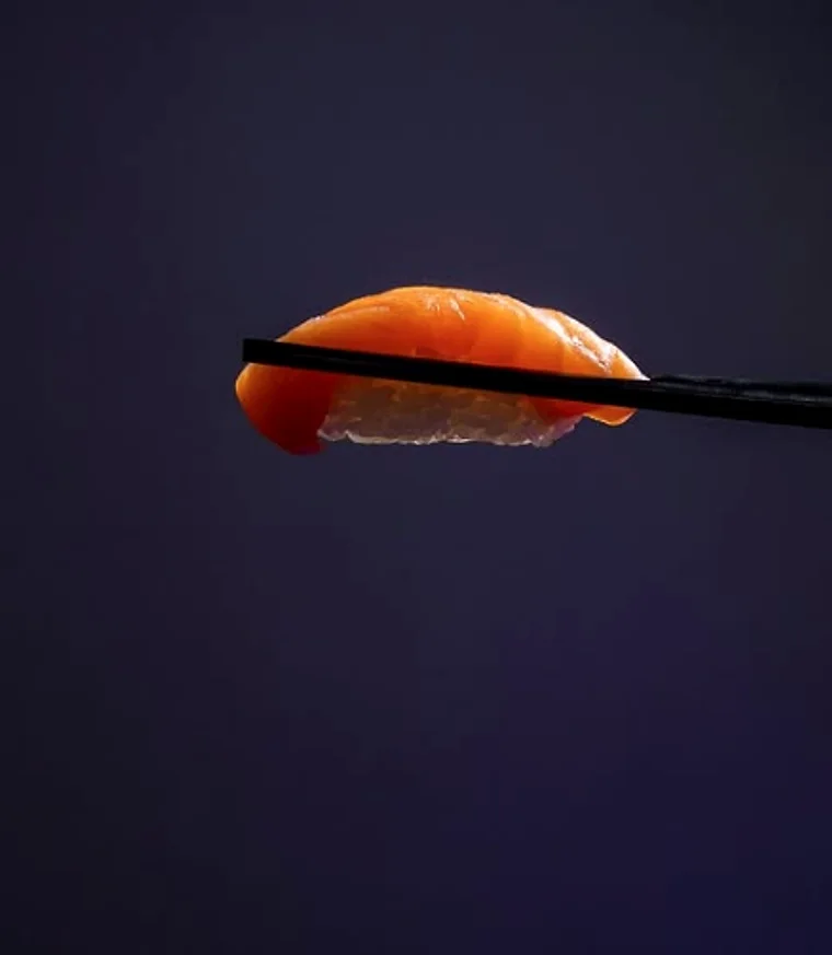 A single piece of salmon nigiri held on chopsticks against a deep blue backdrop
