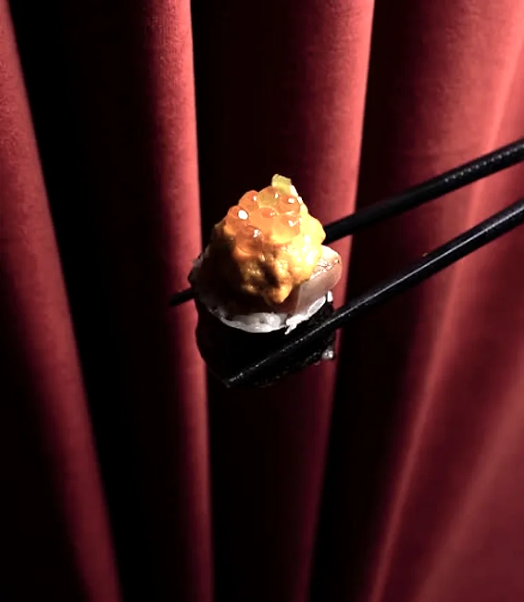A gold-topped bite held on chopsticks against a red velvet curtain