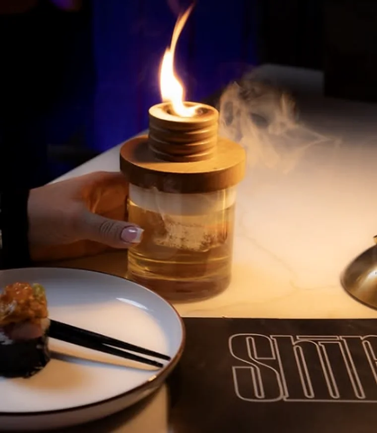 A flame and smoke rising from a wooden vessel beside an etched SHIRO glass