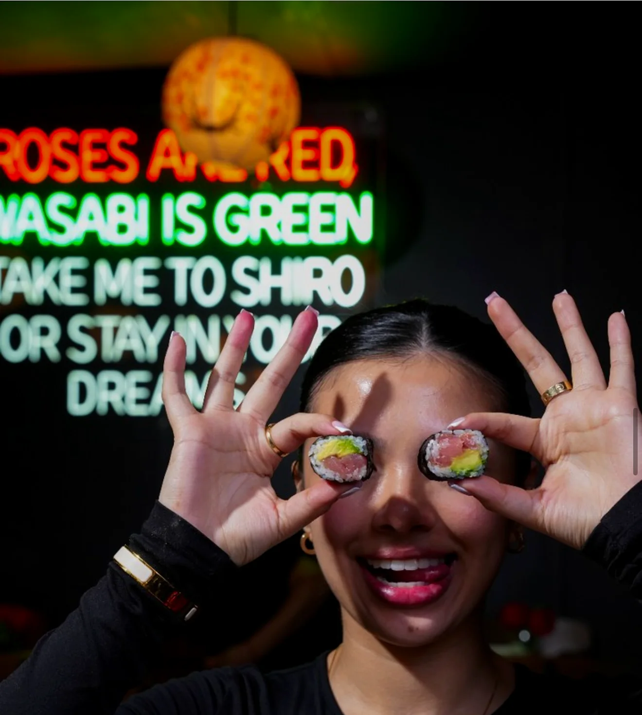 A member of the SHIRO team holding two maki rolls up to her eyes in front of the neon sign reading "Roses are red, wasabi is green, take me to SHIRO or stay in your dreams"
