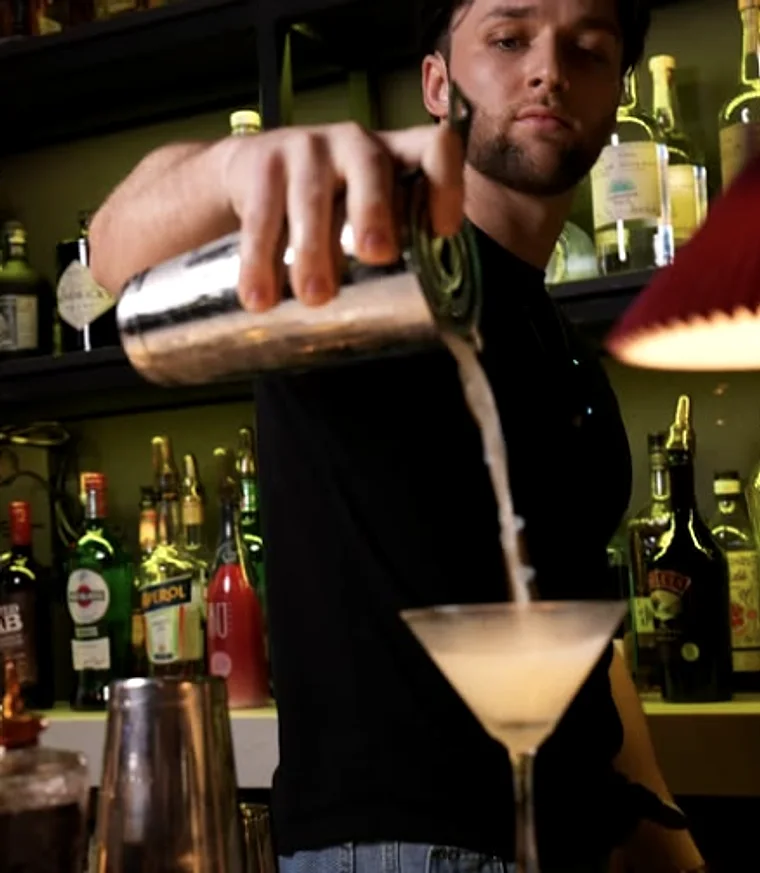 A bartender pouring a cocktail in front of the bottle wall