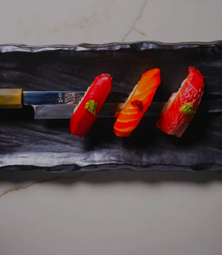 Three pieces of nigiri lined up on slate beside a sushi knife