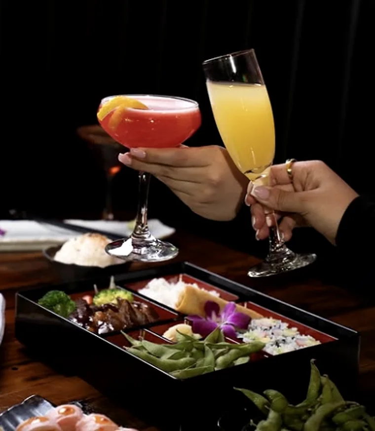 Two hands touching cocktail glasses above a bento of grilled dishes